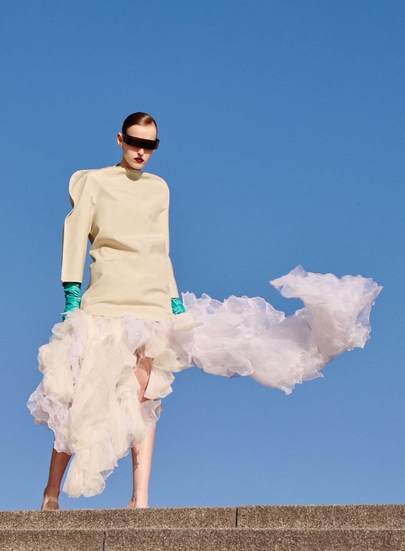 Model wearing pale top and voluminous white ruffled skirt with green gloves against a clear blue sky