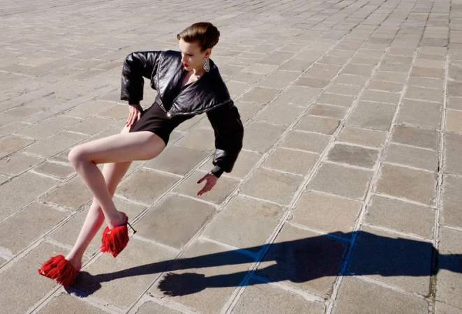 Model in a black leather jacket and red fringed heels posing dynamically on a paved stone public square.