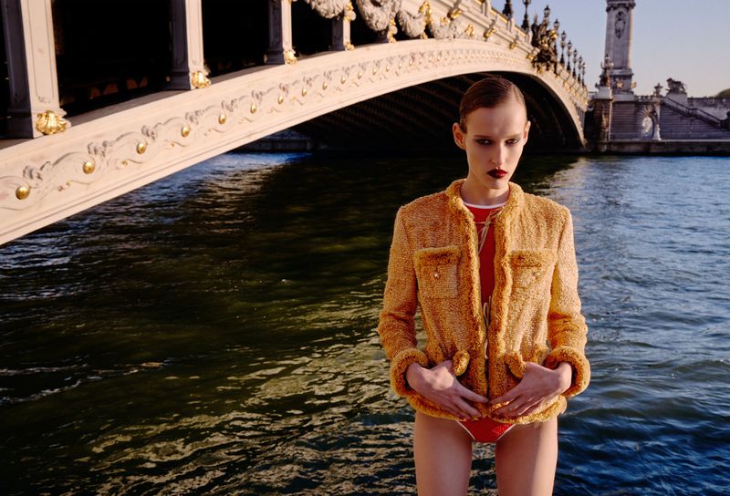 Model wearing a textured orange jacket and red swimsuit standing outdoors by water beneath a stone bridge