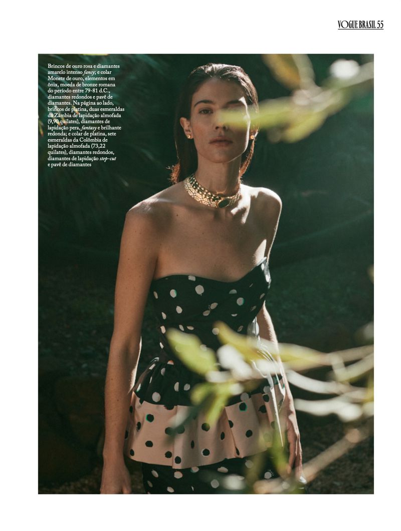 Brunette woman in a black and beige polka-dot dress and a gold necklace poses in a dimly lit natural environment.