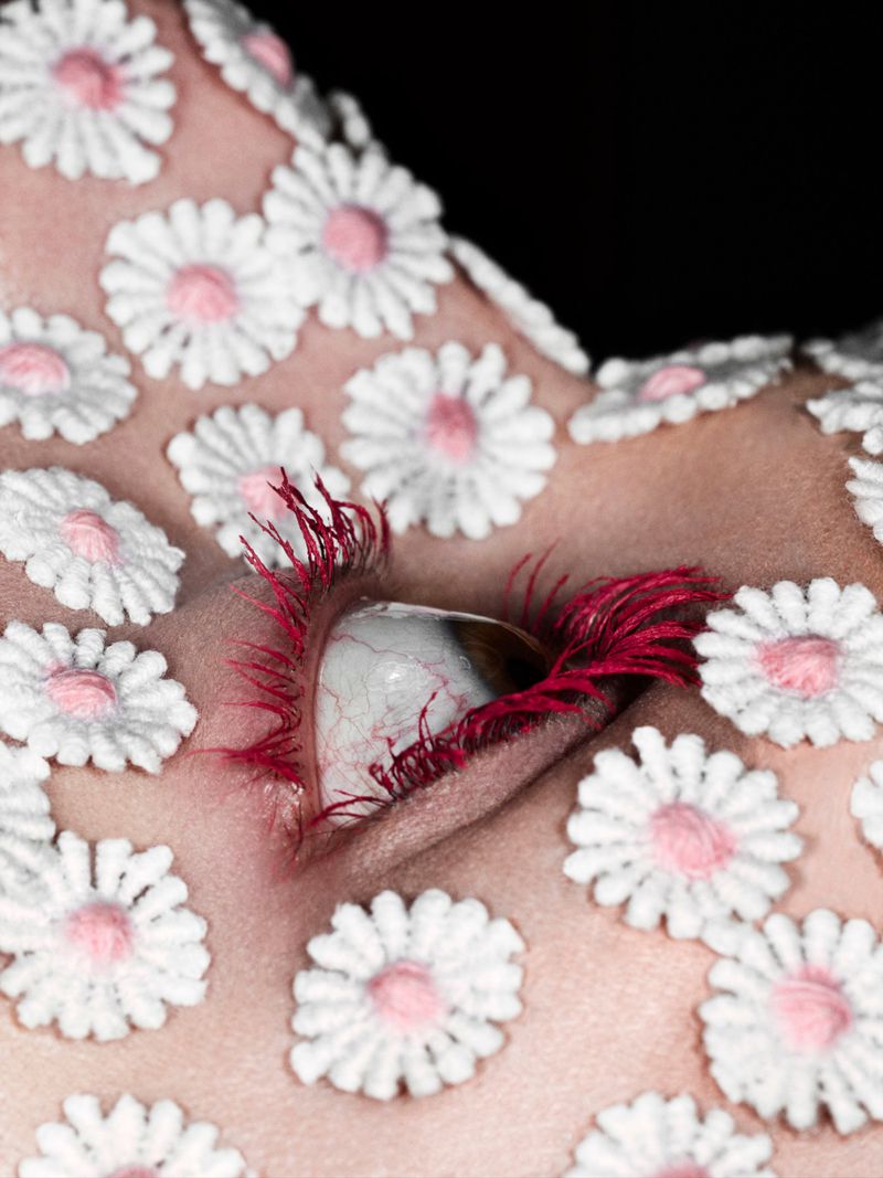 Close-up of a human eye with crimson red eyelashes, surrounded by small embroidered white daisy patches on skin.