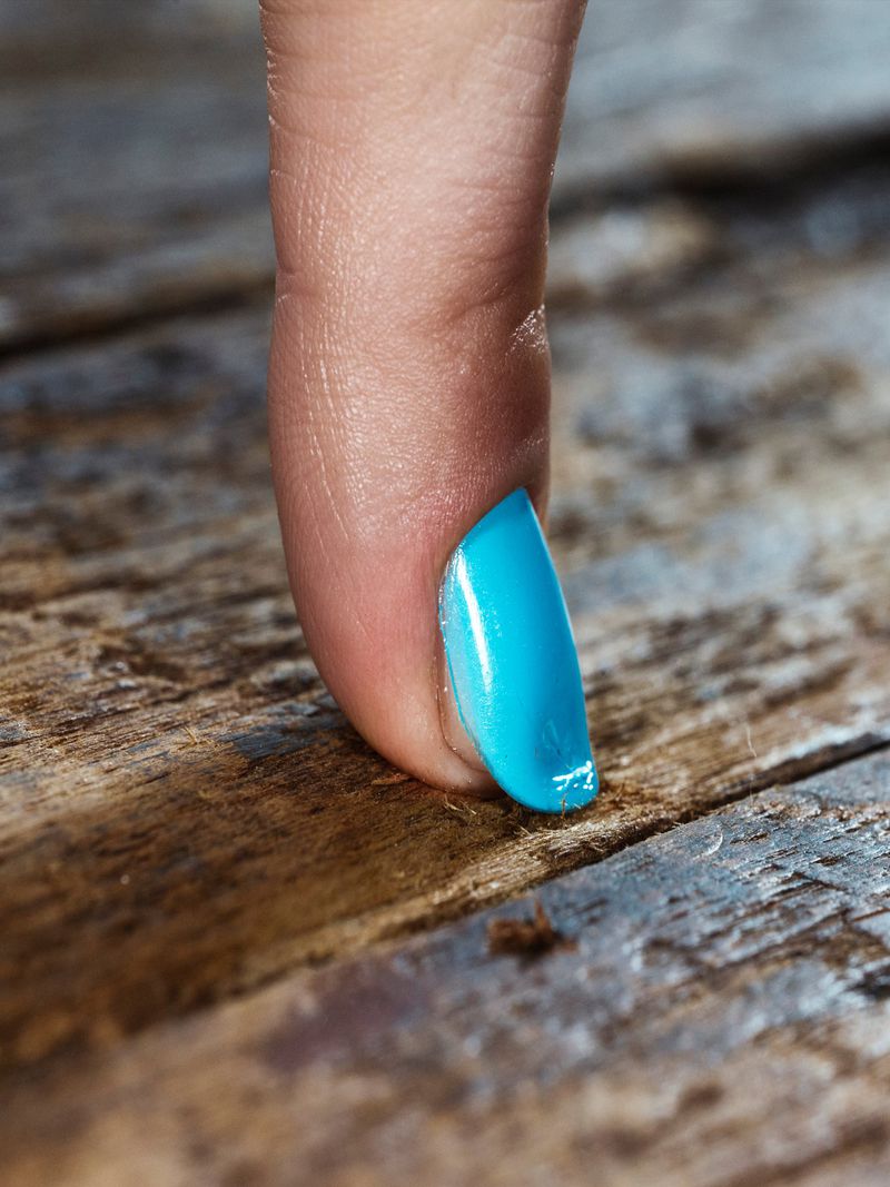 Close-up macro shot of a finger with bright turquoise nail polish touching a weathered wooden surface for Beyond Noise.