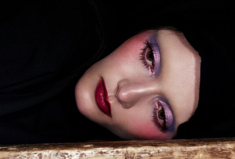 Close-up beauty portrait of a model with dramatic purple eyeshadow and deep red lips against a black background.