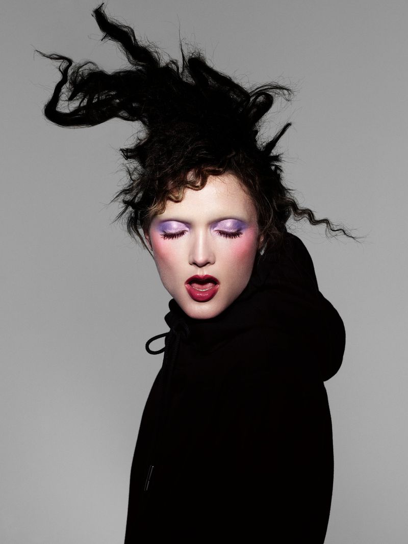 Model with dramatic dark hair and purple eyeshadow wearing a black hoodie against a grey background for Beyond Noise.