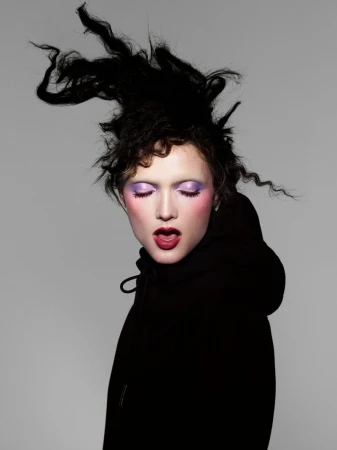 Model with dramatic dark hair and purple eyeshadow wearing a black hoodie against a grey background for Beyond Noise.