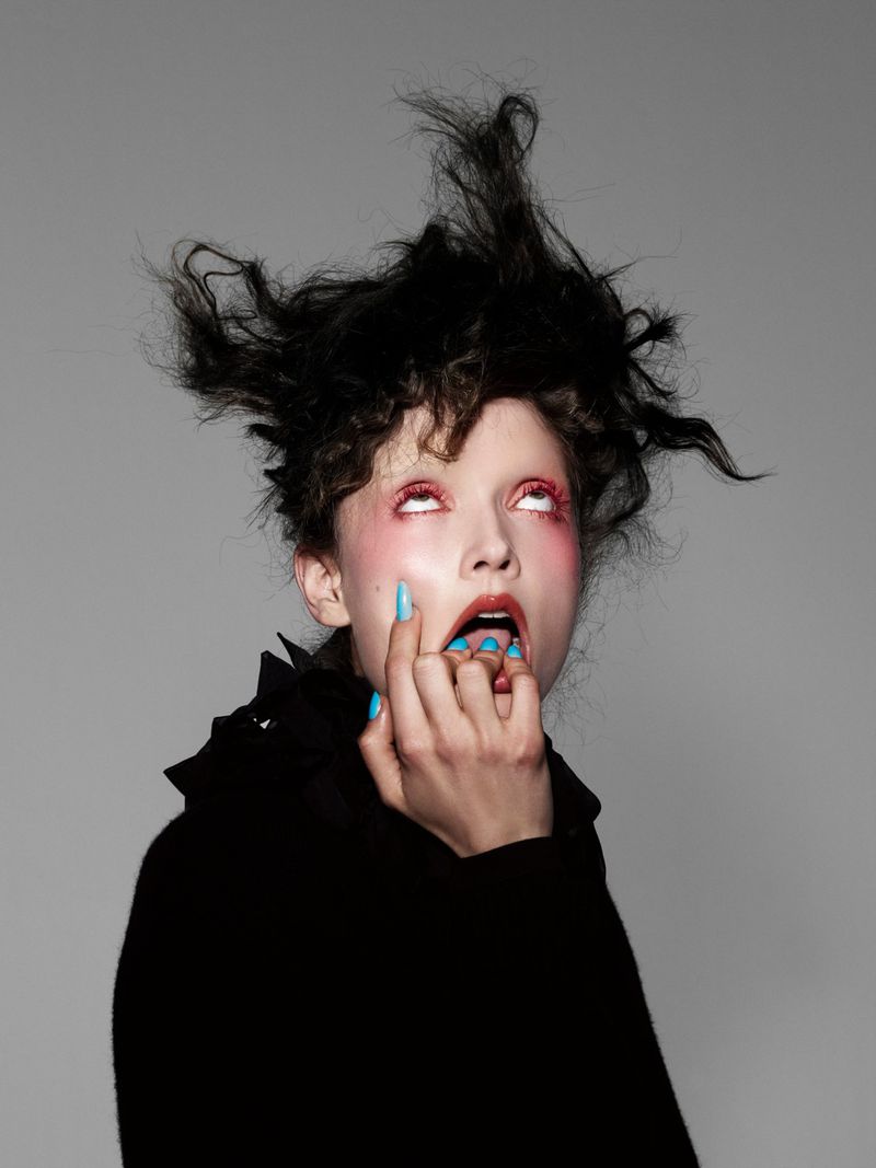 Model with wild dark hair and blue nails biting fingers against a grey background