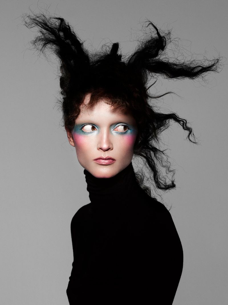 Model with avant-garde teased black hair and blue and pink eye makeup wearing a black turtleneck against a neutral grey background