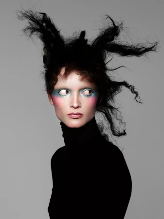 Model with avant-garde teased black hair and blue and pink eye makeup wearing a black turtleneck against a neutral grey background