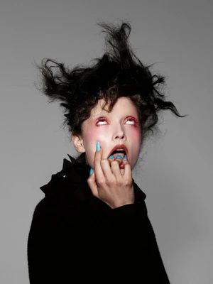 Model with wild dark hair and blue nails biting fingers against a grey background