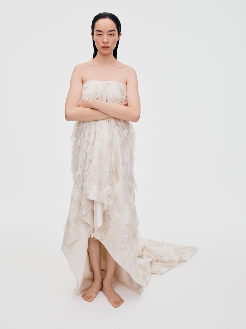Full-length portrait of an Asian woman posing in a light strapless feathered gown in a white studio with her arms crossed.