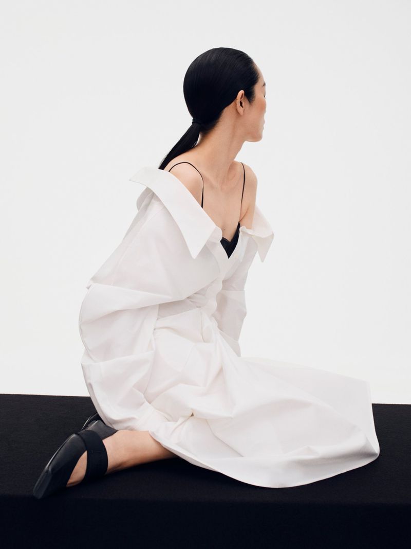 Model Feifei Sun seated in a white oversized shirt and black shoes against a white background for Beyond Noise