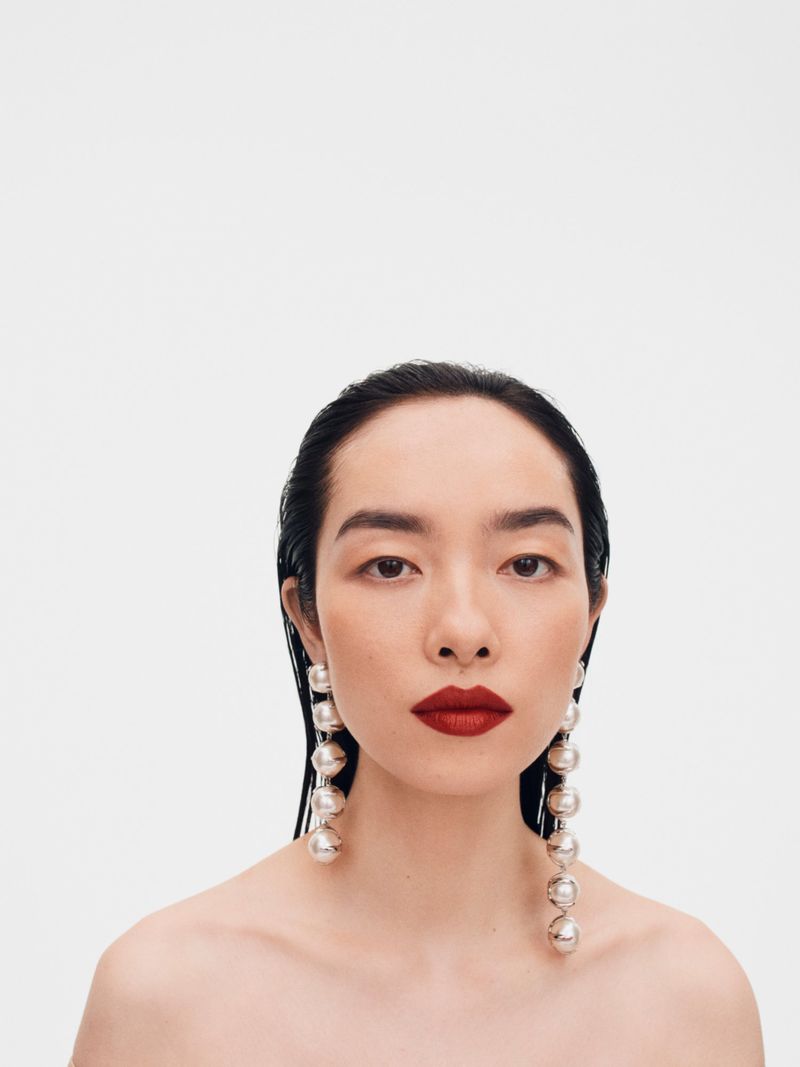 Portrait of Feifei Sun wearing long pearl drop earrings and red lipstick against a white seamless backdrop