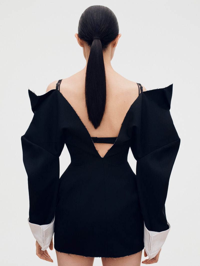Model with long dark ponytail seen from behind wearing a black structured dress with white cuffs against white seamless background