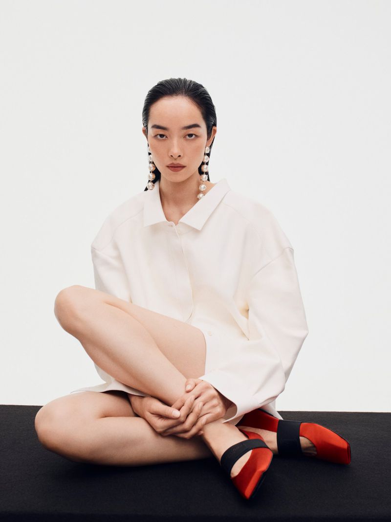 Model FeiFei Sun seated on a black surface against a white wall wearing a white shirt and red shoes