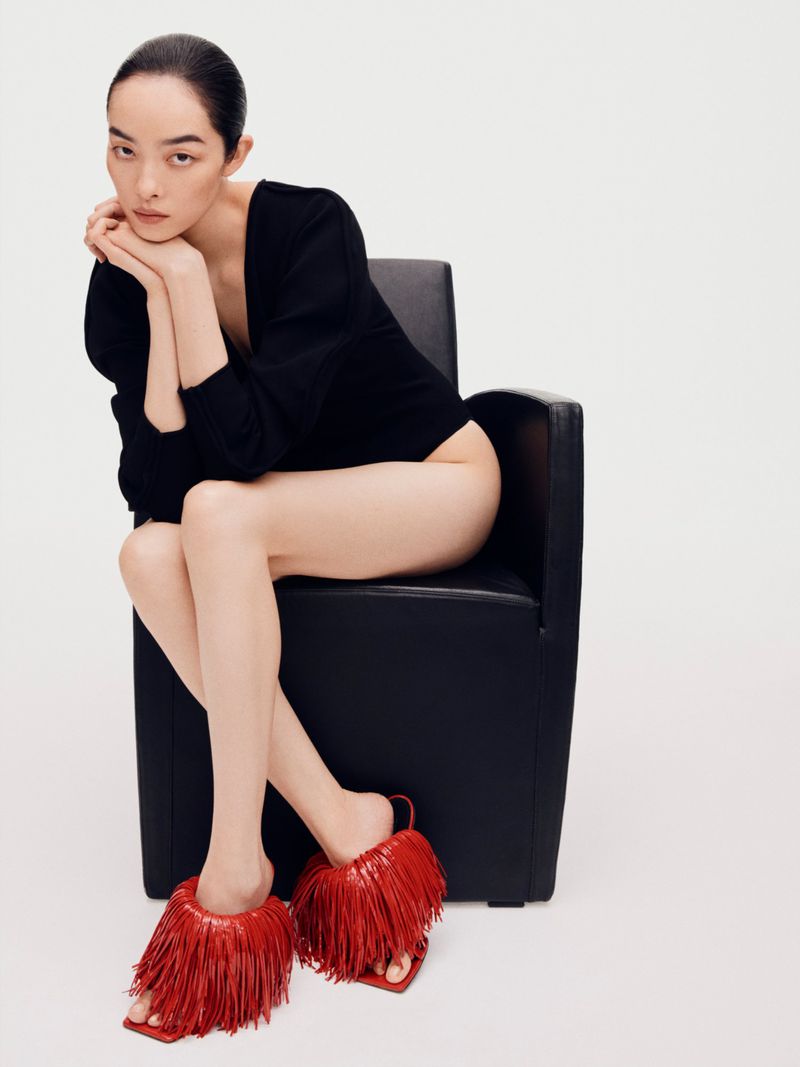 Model Feifei Sun seated in a black armchair wearing a black top and red fringed mules against a white background