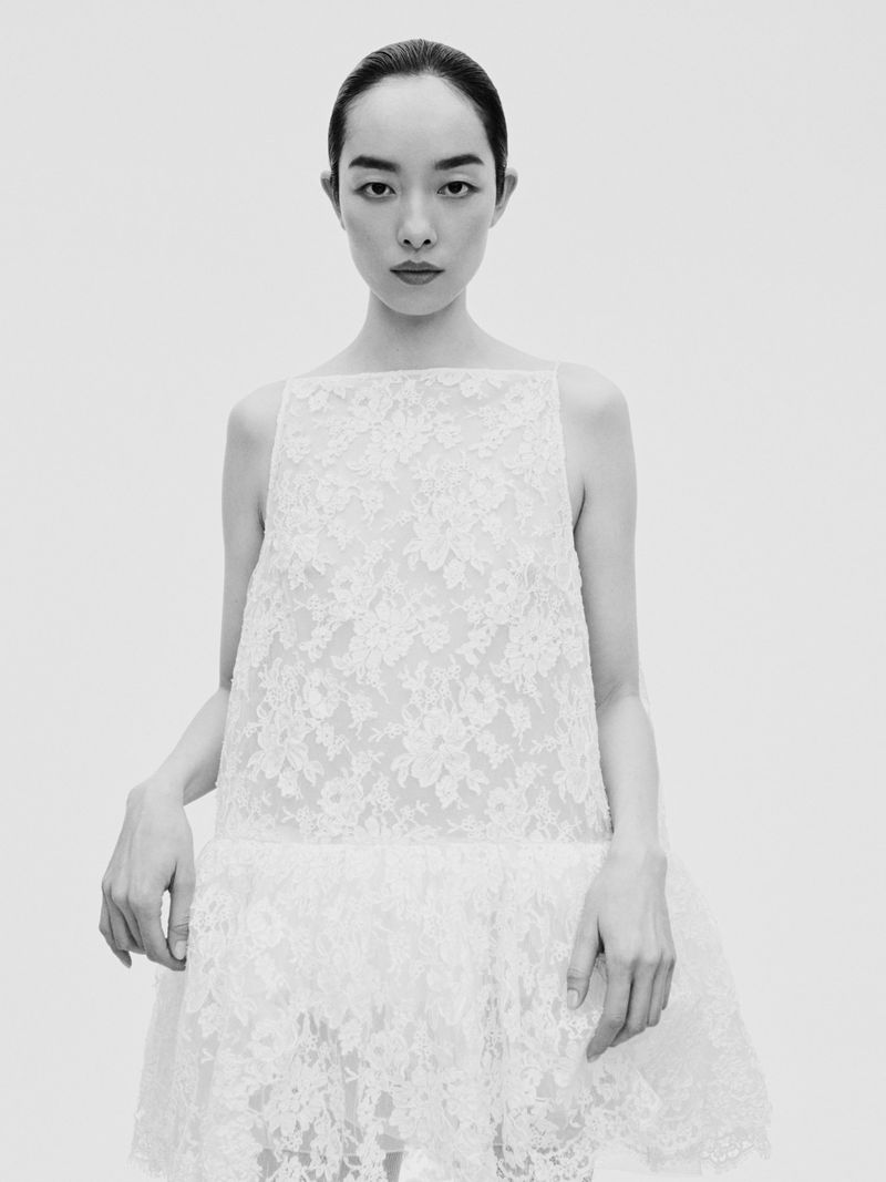 Model FeiFei Sun wearing a white lace dress against a neutral seamless studio backdrop in an editorial portrait.