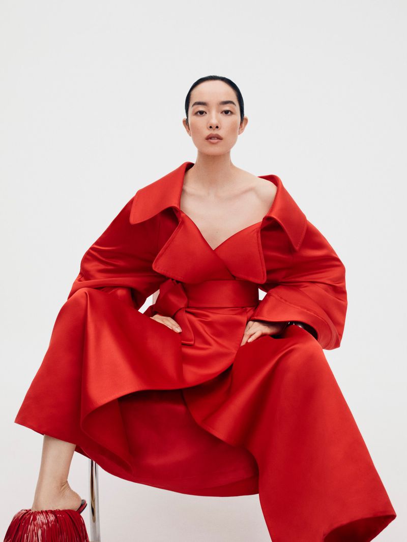 Model FeiFei Sun wearing a voluminous red silk coat and matching red mules, seated against a white seamless backdrop.