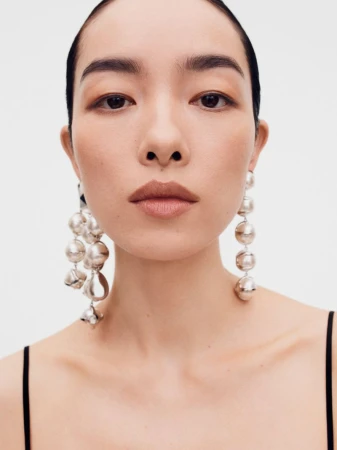 Close-up portrait of model FeiFei Sun wearing large metallic statement earrings against a plain white background