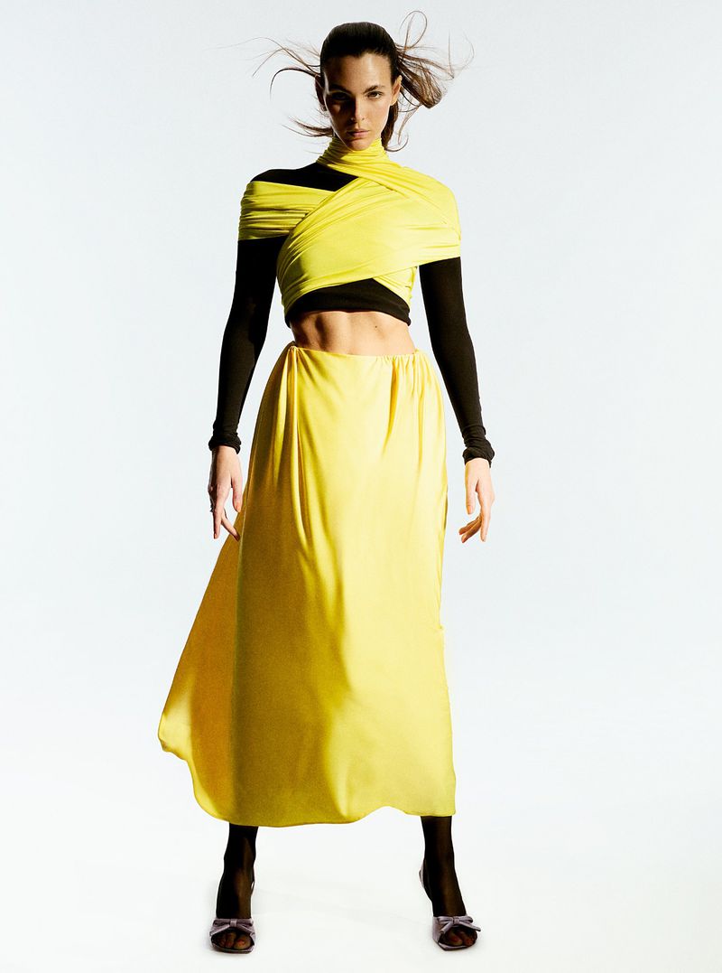A woman in a yellow skirt and top poses in a full shot. The background is a light color. Her hair is long and flowing.