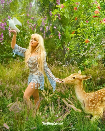 Woman with long blonde hair stands in a grassy field with a fawn, holding a white dove, surrounded by flowers and greenery.