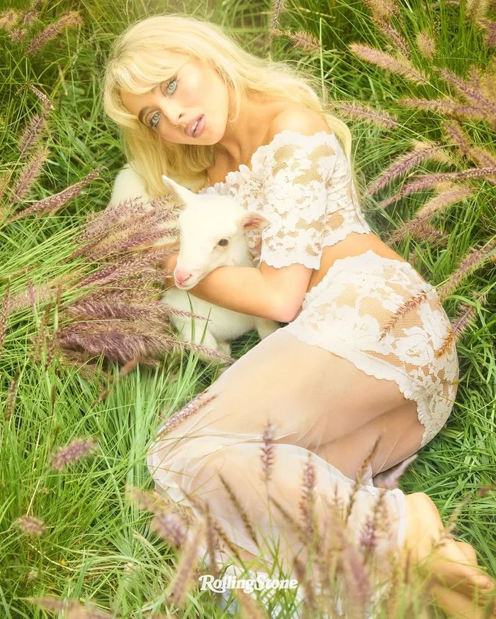 Sabrina Carpenter lying in a grassy meadow holding a small white lamb, wearing a lace two-piece outfit.