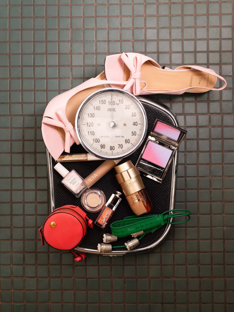 Scale surrounded by beauty products including blush, lip gloss, foundation, nail polish and shoes on a tiled surface.