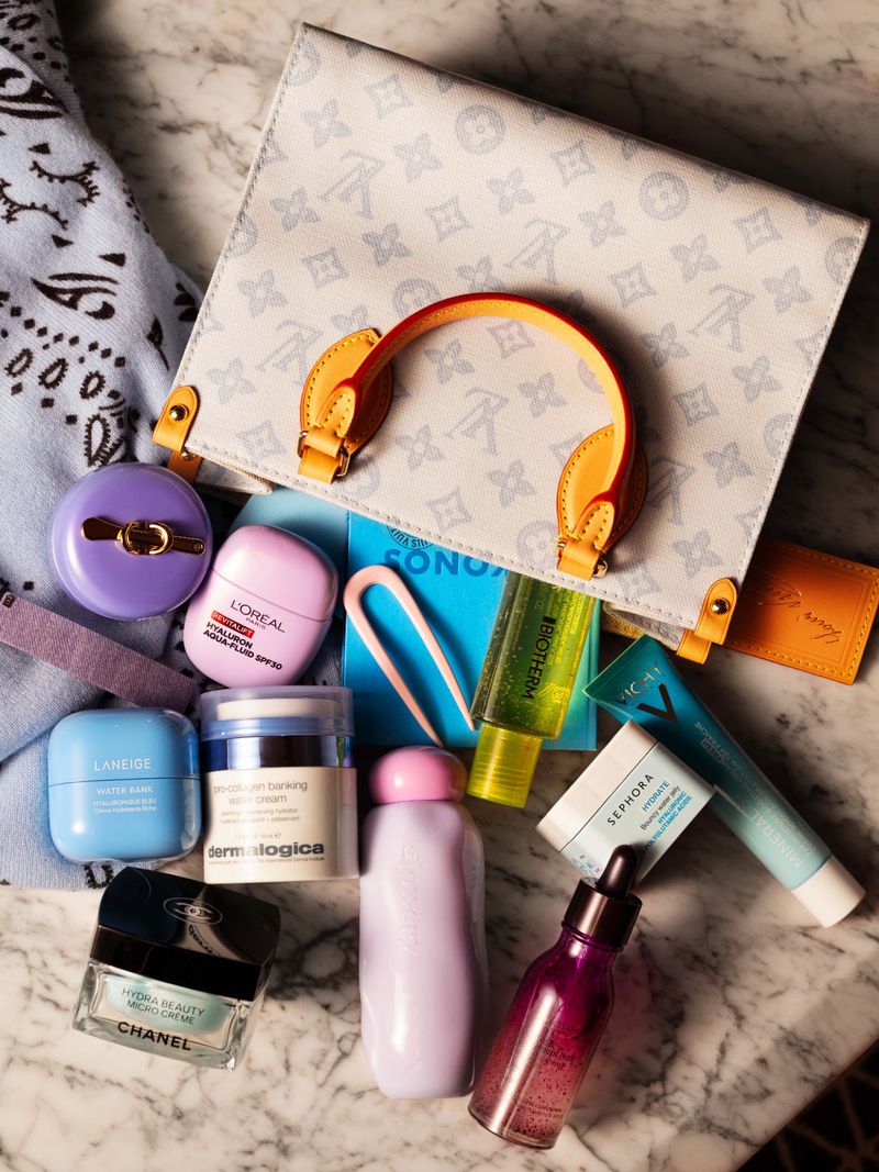 Flat lay of luxury designer handbag and beauty products on a white marble surface for Grazia France magazine.