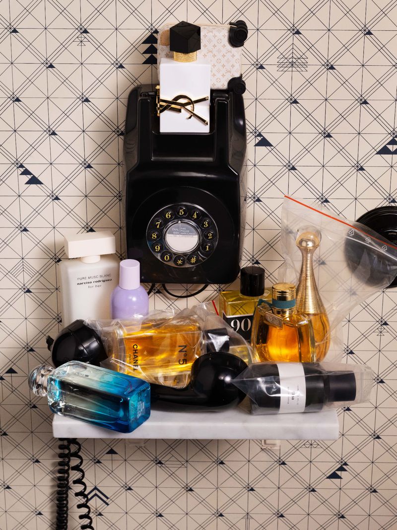 Collection of perfume bottles and a vintage black rotary telephone displayed on a small marble shelf against patterned wallpaper