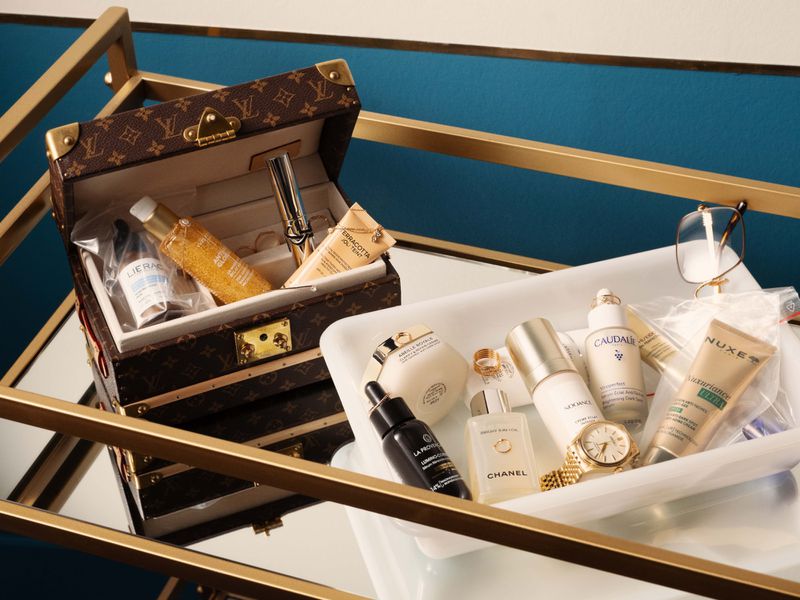 Beauty products and skincare bottles arranged in a white ceramic tray beside a monogrammed vanity case on a glass-topped gold metal cart.
