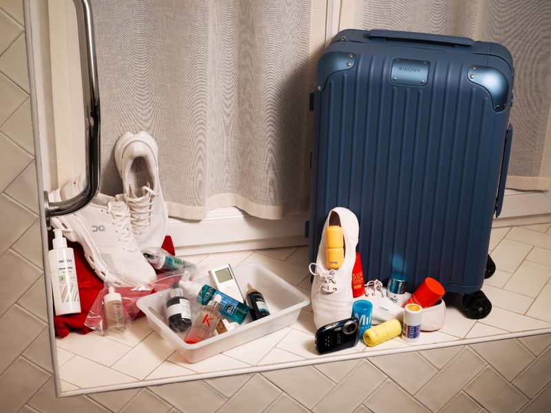 Blue suitcase next to white sneakers and travel toiletries arranged on a tiled floor for Grazia France editorial.