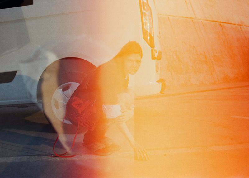 Crouching woman in dark clothing near a white vehicle with a strong orange and red light leak across the frame.