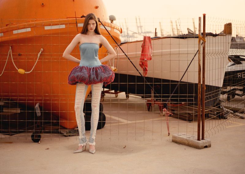 Model wearing a strapless blue top and ruffled skirt with distressed leggings standing before a large orange lifeboat in a port
