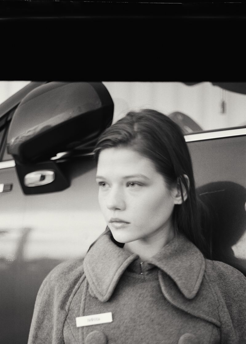 Monochrome portrait of a young woman wearing a buttoned coat leaning against a car exterior.