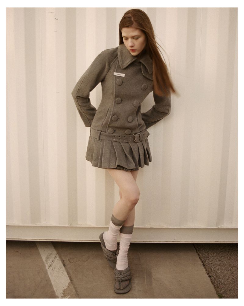 A young female model wearing a fitted grey double-breasted coat and pleated skirt standing against a vertical paneled background