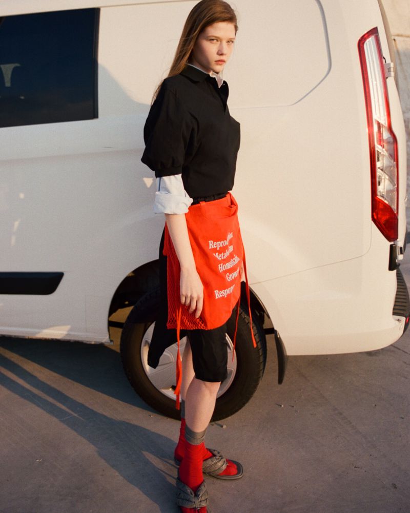 Young woman standing beside a white van wearing a black polo shirt, layered white cuffs, red skirt wrap, and red socks.