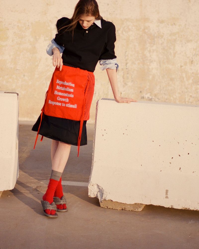 Model standing outdoors wearing a black polo shirt, red skirt with white text, red socks, and grey slides, leaning on a concrete barrier