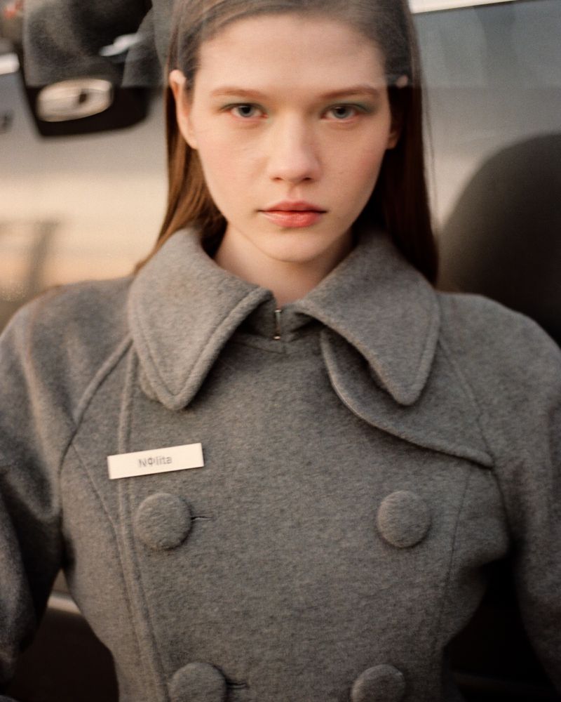 Portrait of a young woman wearing a grey double-breasted wool coat with a name tag, outdoors against a vehicle background.