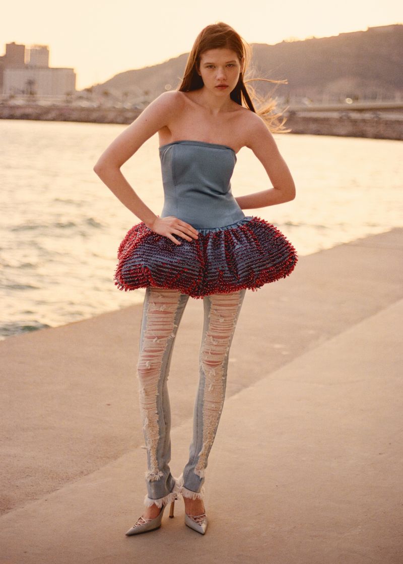 Woman in blue top, red skirt, and ripped jeans poses on a waterfront promenade with buildings and a mountain in the background.