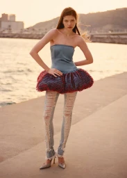 Model standing on a waterfront promenade wearing a blue strapless top, red textured skirt, and distressed jeans