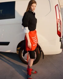 Young woman standing beside a white van wearing a black polo shirt, layered white cuffs, red skirt wrap, and red socks.