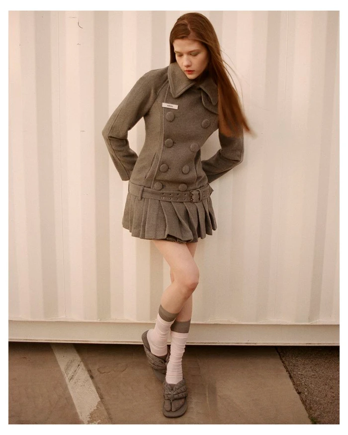 A young female model wearing a fitted grey double-breasted coat and pleated skirt standing against a vertical paneled background