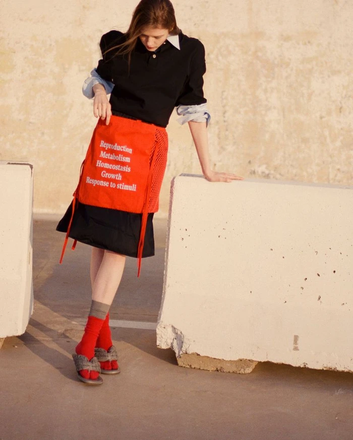 Model standing outdoors wearing a black polo shirt, red skirt with white text, red socks, and grey slides, leaning on a concrete barrier