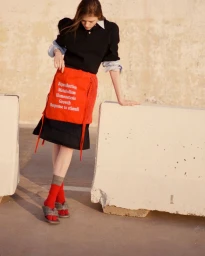 Model standing outdoors wearing a black polo shirt, red skirt with white text, red socks, and grey slides, leaning on a concrete barrier