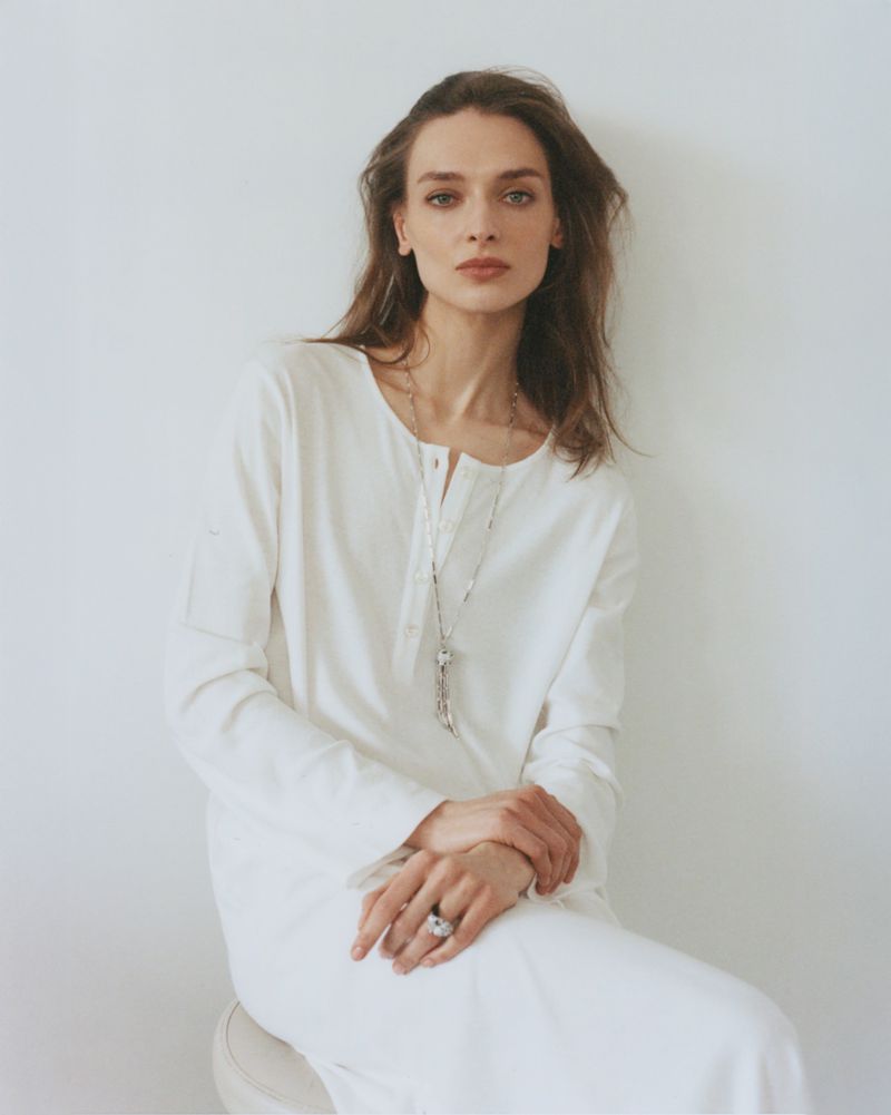 Portrait of a young adult woman with long brown hair, wearing a white linen dress, sitting against a white wall in a natural light setting.