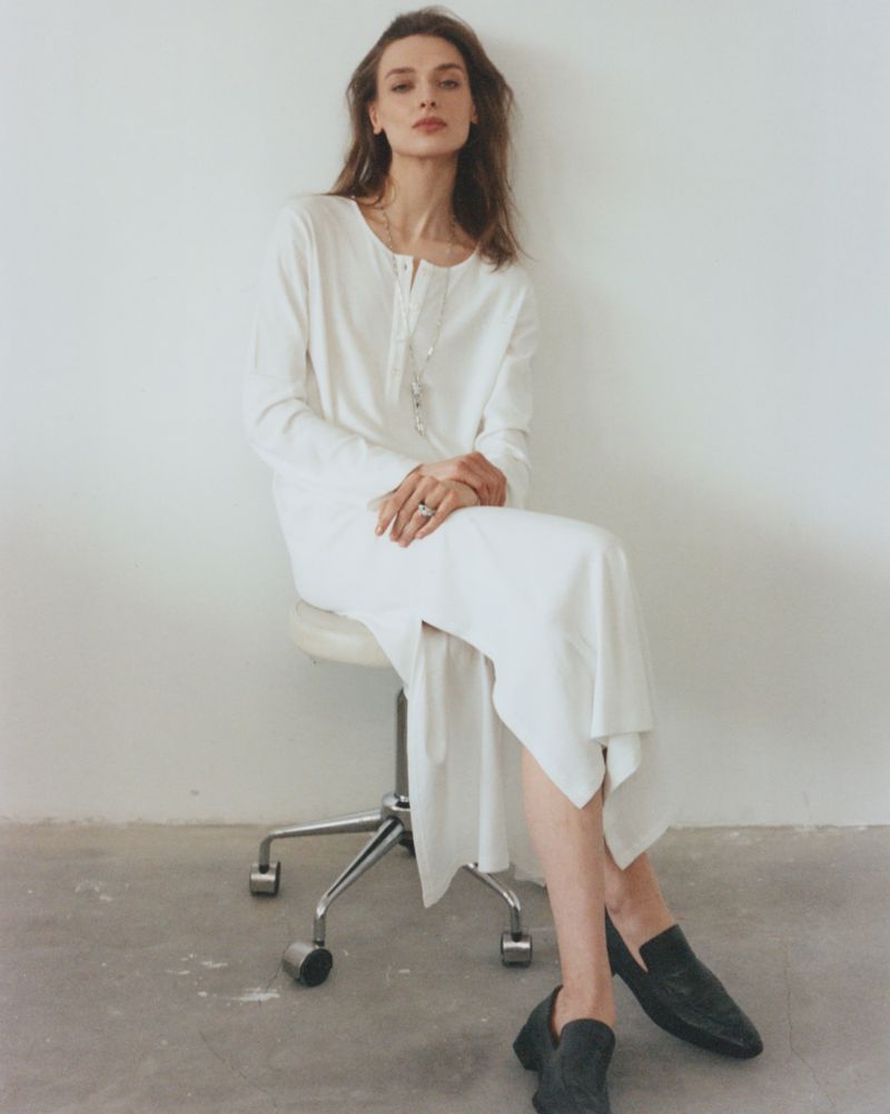 Model Daga Ziober seated on a stool wearing a long white dress and black loafers against a seamless white background