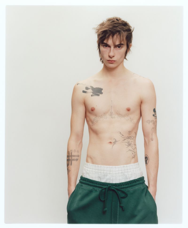 Shirtless young man with tattoos wearing green sweatpants against a plain white studio background