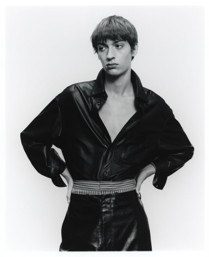 Model wearing a black leather shirt and trousers with hands on hips against a white studio background