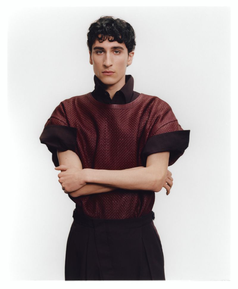 Portrait of a man wearing a textured burgundy short-sleeve top with dark trousers against a white backdrop.