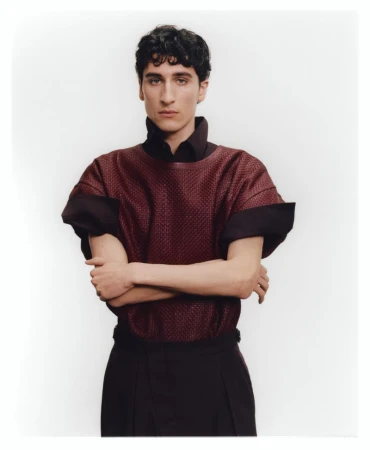 Portrait of a man wearing a textured burgundy short-sleeve top with dark trousers against a white backdrop.