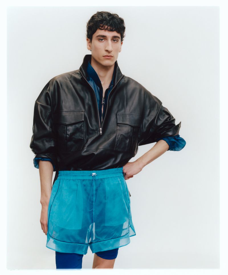 Male fashion model poses in a photo studio wearing a black leather jacket and turquoise mesh shorts in a medium shot.